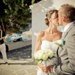 A newly wed couple kiss in front of a Greek chapel. In the background you can see the sea and Mark Usher, Musician and Wedding DJ in Rhodes, Greece playing the guitar.