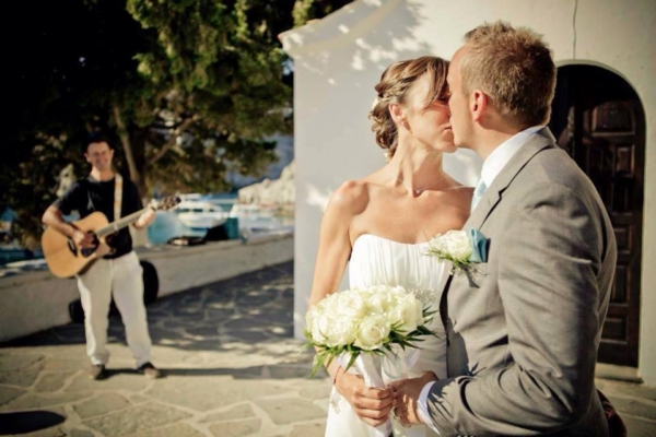 A newly wed couple kiss in front of a Greek chapel. In the background you can see the sea and Mark Usher, Musician and Wedding DJ in Rhodes, Greece playing the guitar.