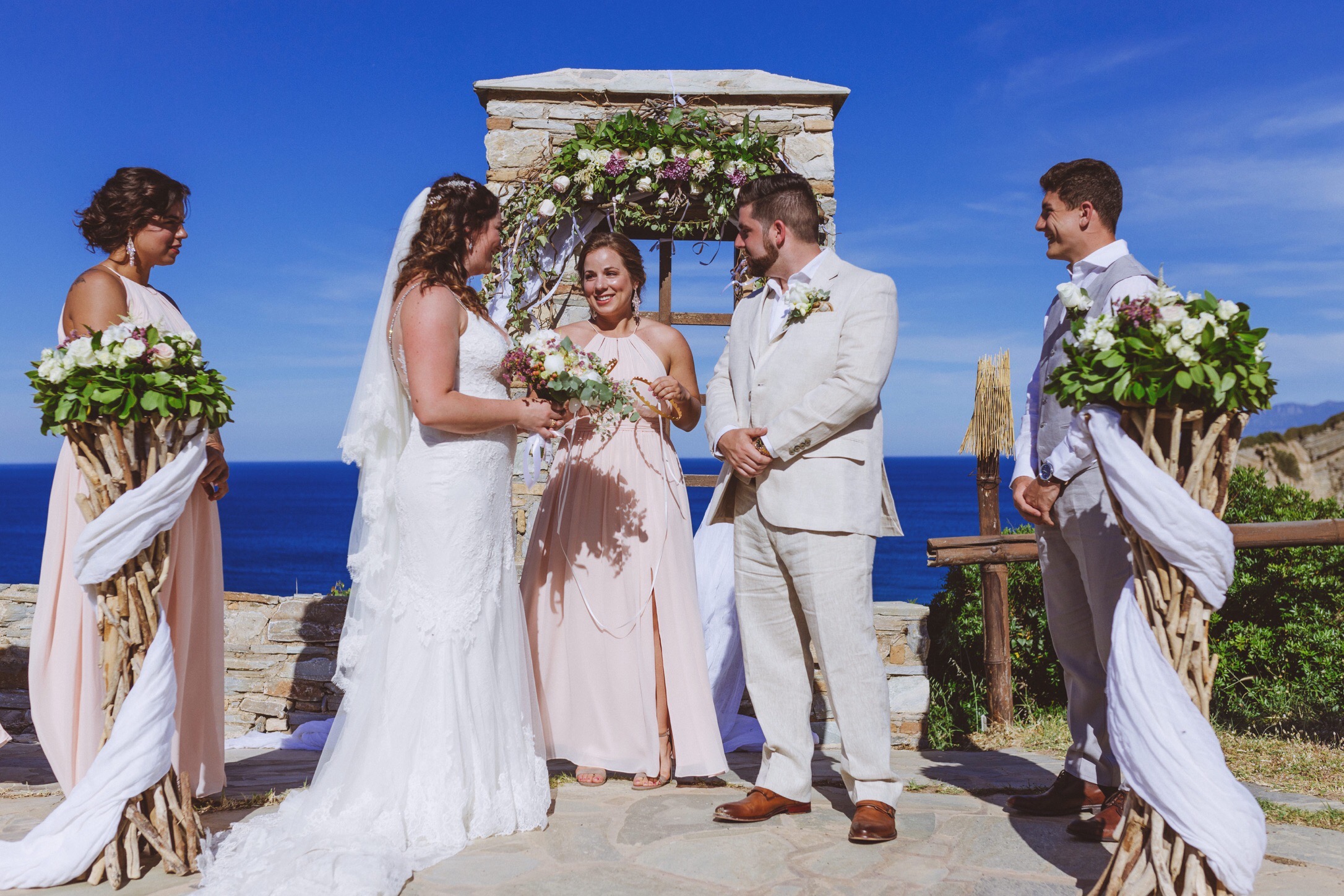 A Greek island wedding at Villa Delenia on the island of Evia, Greece. The couple are standing in front of blue sea and sky whilst a bridesmaid prepares to place Stefania, or Greek wedding crowns