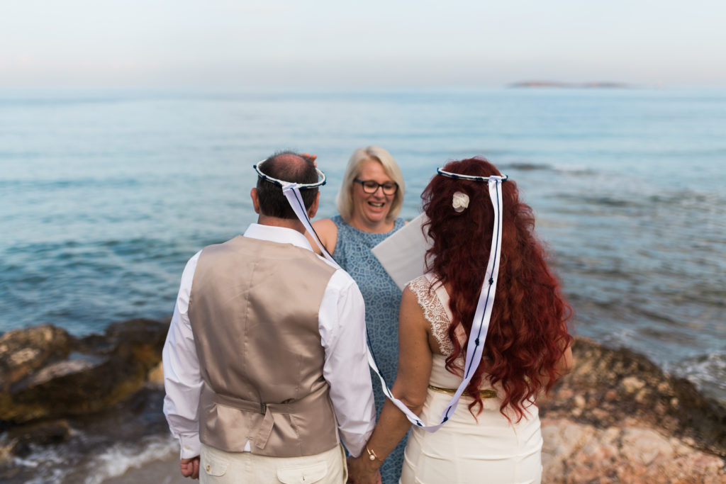 a couple face the sea wearing greek wedding corwns as they exchange their wedding vows with a celebrant in greece