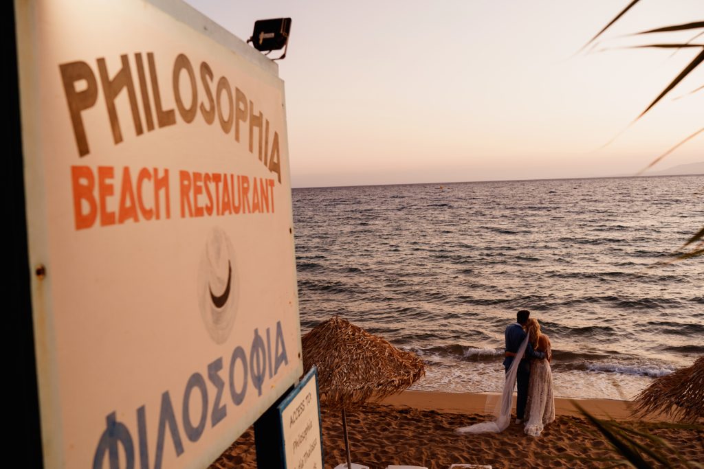 A sign saying 'Philosophia beach restaurant with a newly married couple standing on the beach looking out to sea after their wedding ceremony at Philosophia with ECK A wedding Celebrant in Greece