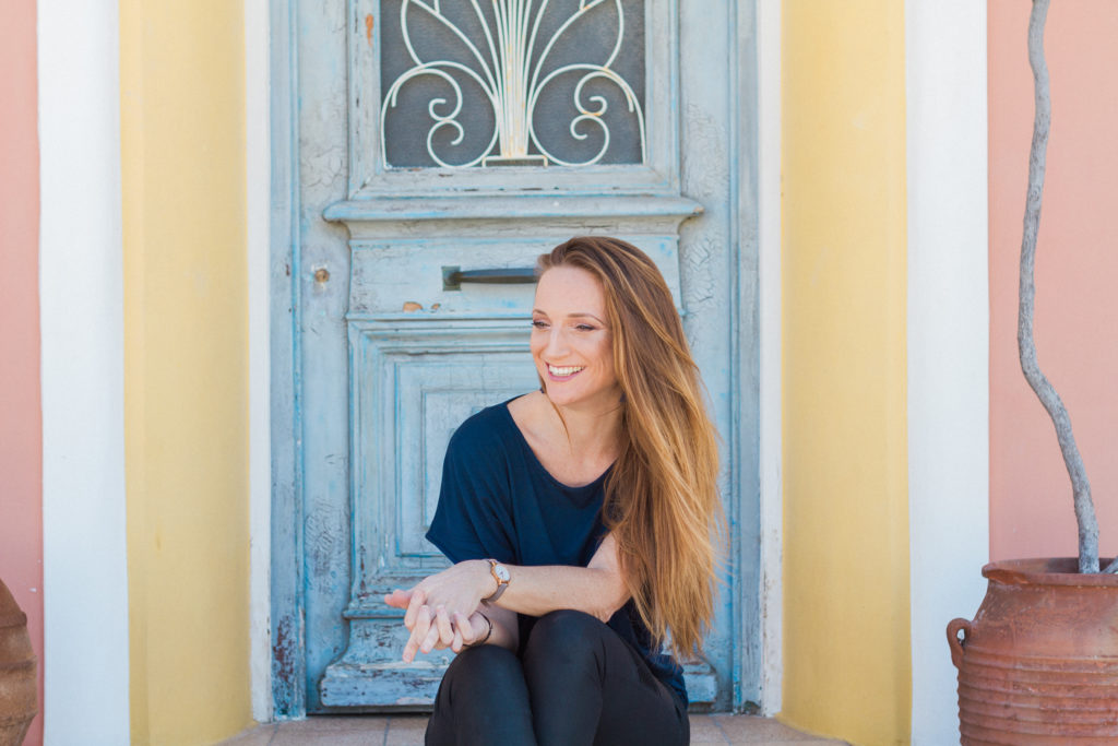 a photo of a young woman with long hair sitting in a doorway with Greek pastel colours. This is Claire from Lefkas weddings, she can design your wedding in Lefkas for you!