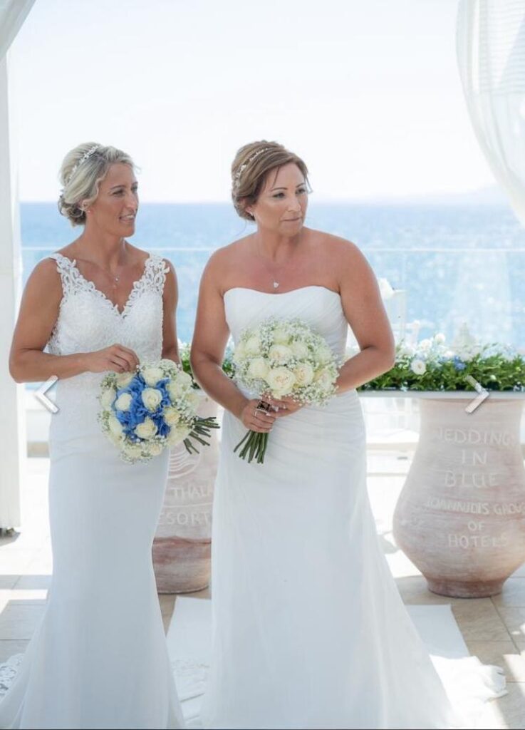A lesbian wedding in Greece, the couple hold their bouquets and are standing in front of the blue sea.