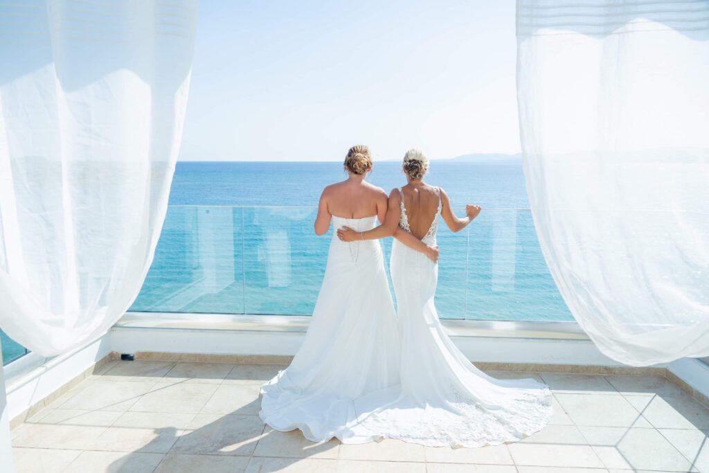A lesbian wedding couple enjoy the view from the veranda at Pefki Blue Hotel on their wedding day. The couple are wearing white wedding dresses and are looking out over the blue sea,