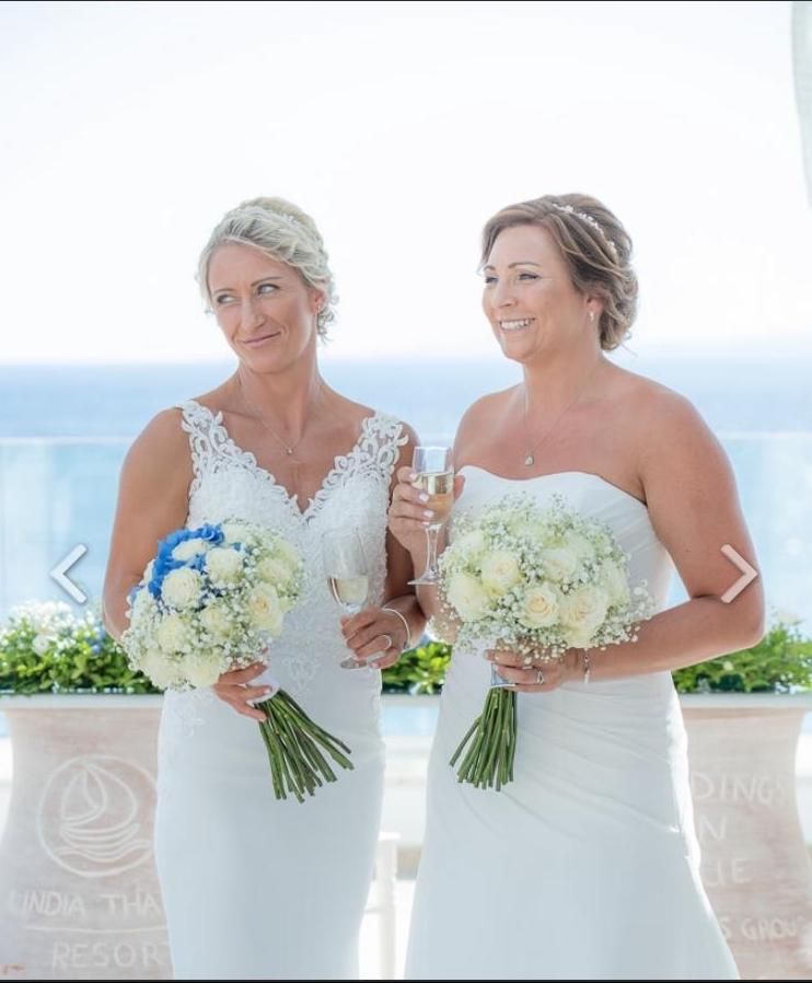 A lesbian wedding in Greece, the couple are holding bouquets and the blue sea is behind them.