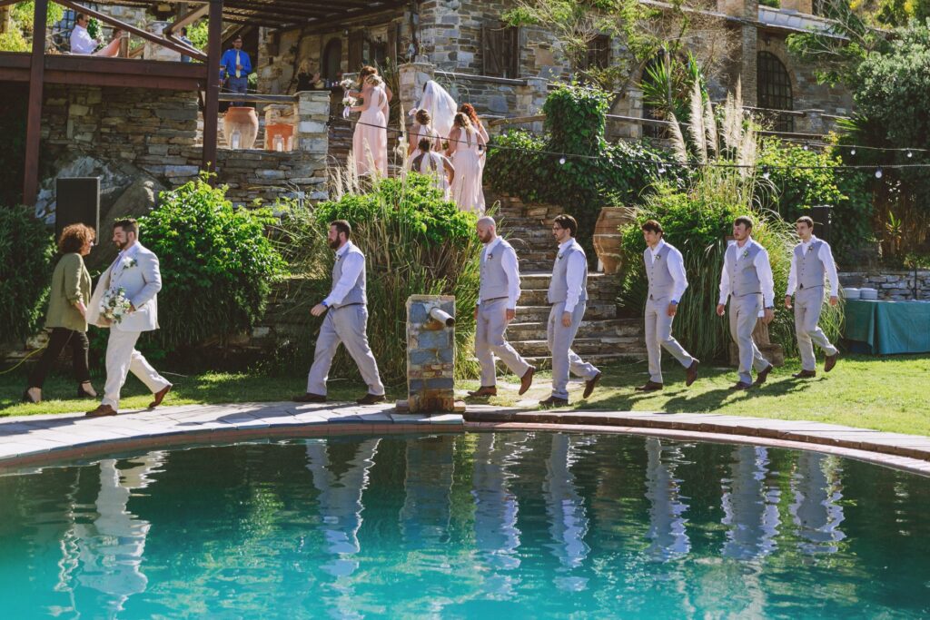 The groom and six groomsmen walk to their places for a wedding ceremony at Villa Delenia in Evia, Greece. They are reflected in the swimming pool.