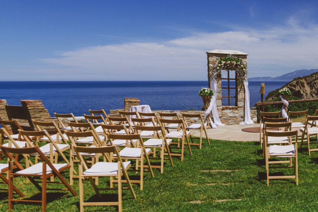 A wedding ceremony set up at the Villa Delenia in Evia, Greece. Simple wooden chairs look forward to a decorative stone window frame with a view to the blue sea beyond.