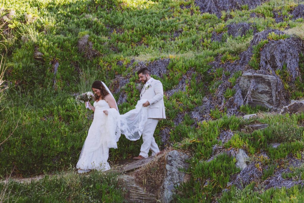 The wedding couple walk down a path surrounded by greenery and rocks. The groom is behind and holding the long veil. The location is Villa Delenia on the greek island of Evia.