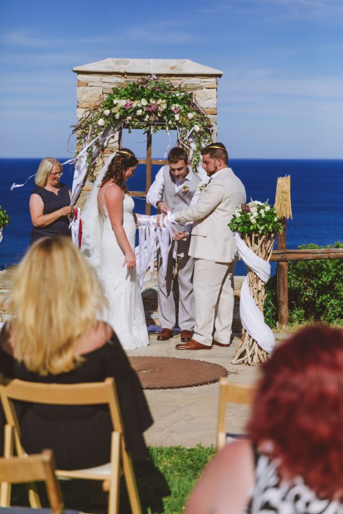 A grromsman ties a hand fasting ribbon, of which there are already many, around the clasped hands of the couple. The wedding celebrant stands to the side and in the background is the beautiful sea view from Villa Delenia