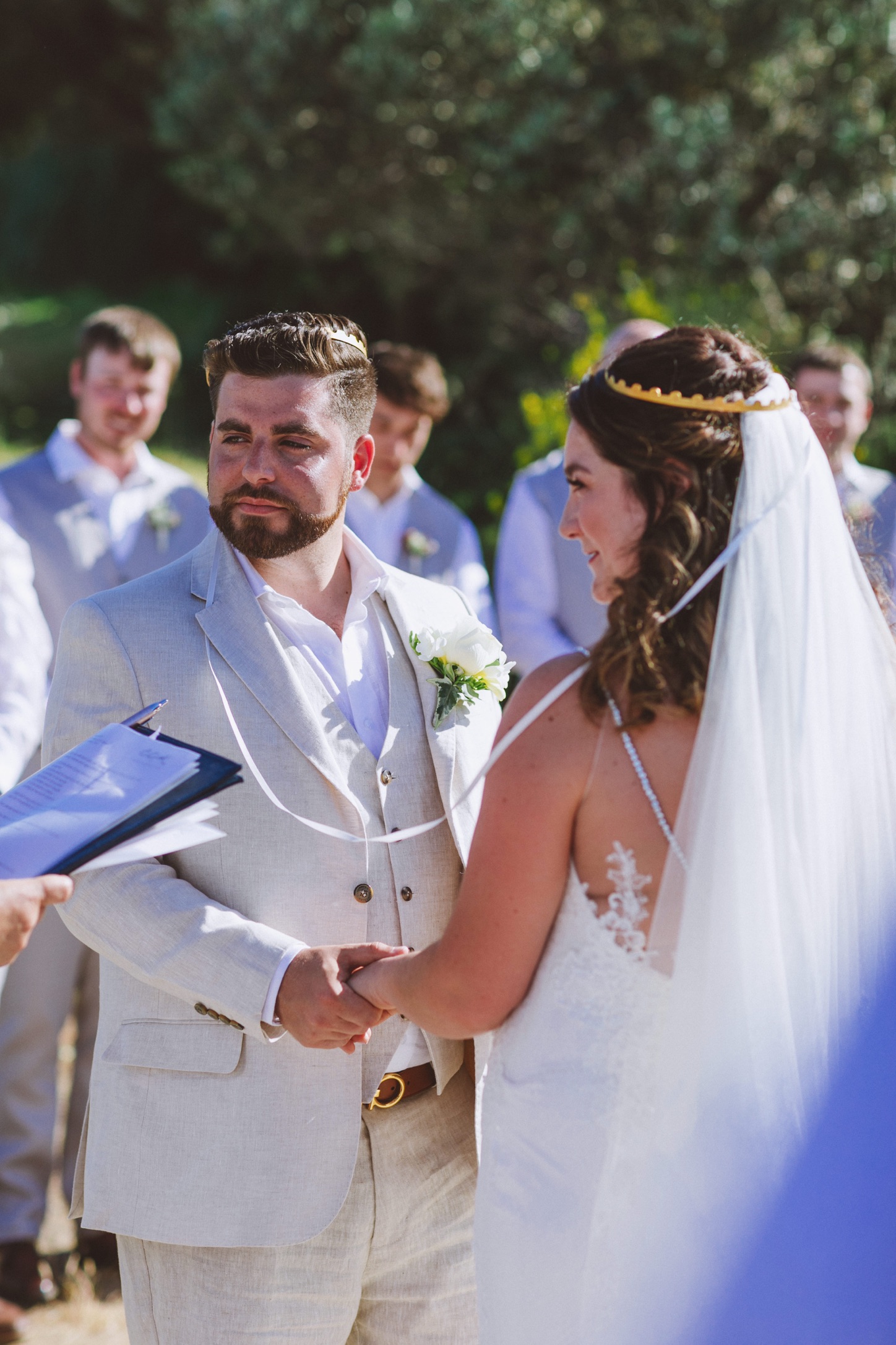 A bride and groom hold hands as they wear the greek wedding crowns and look to the wedding celebrant as their vows are read to them. The ceremony is at Villa Delenia in Evia, Greece